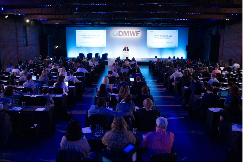 A photo of the Global DMWF conference with attendees seated in rows and a speaker on stage.